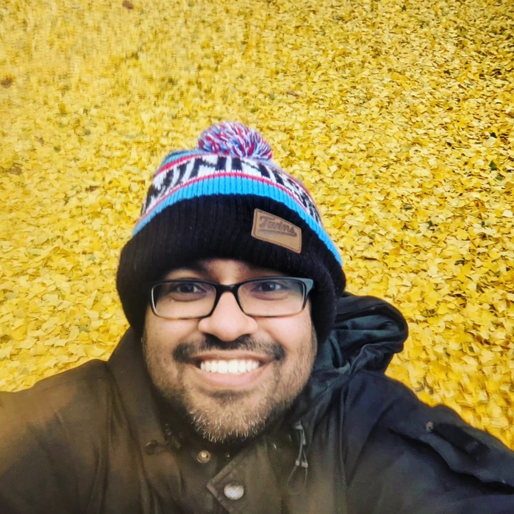 Picture of a man with a beanie on in front of a pile of yellow leaves.