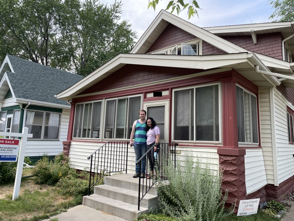 A young couple posing in front of their craftsman-style home in the Midway with red and white accents.